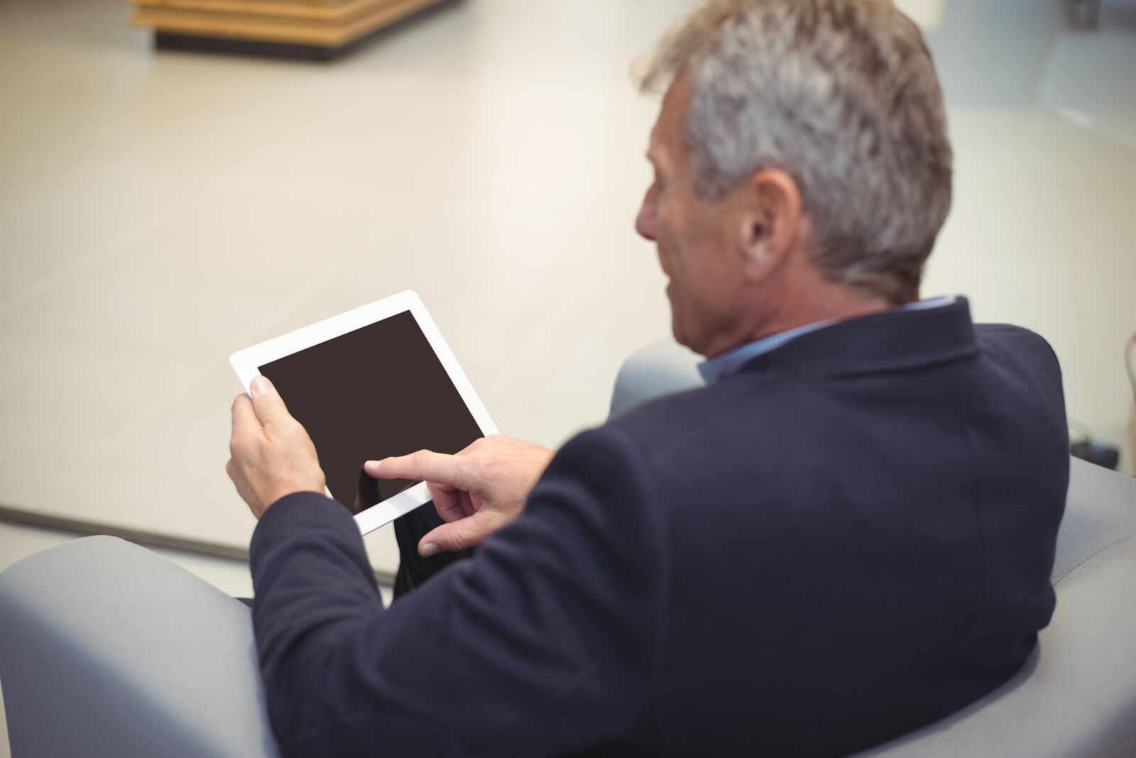 Stariji muškarac u odelu sedi u fotelji i koristi beli tablet uređaj