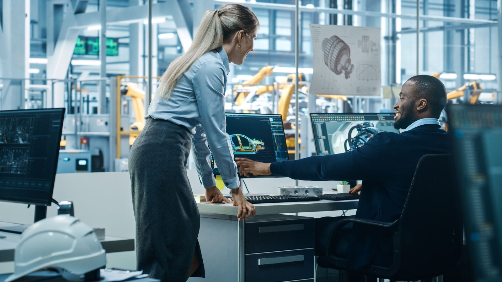 Zaposleni u kancelariji razgovaraju ispred monitora sa tehničkim nacrtima u svetloj industrijskoj hali.