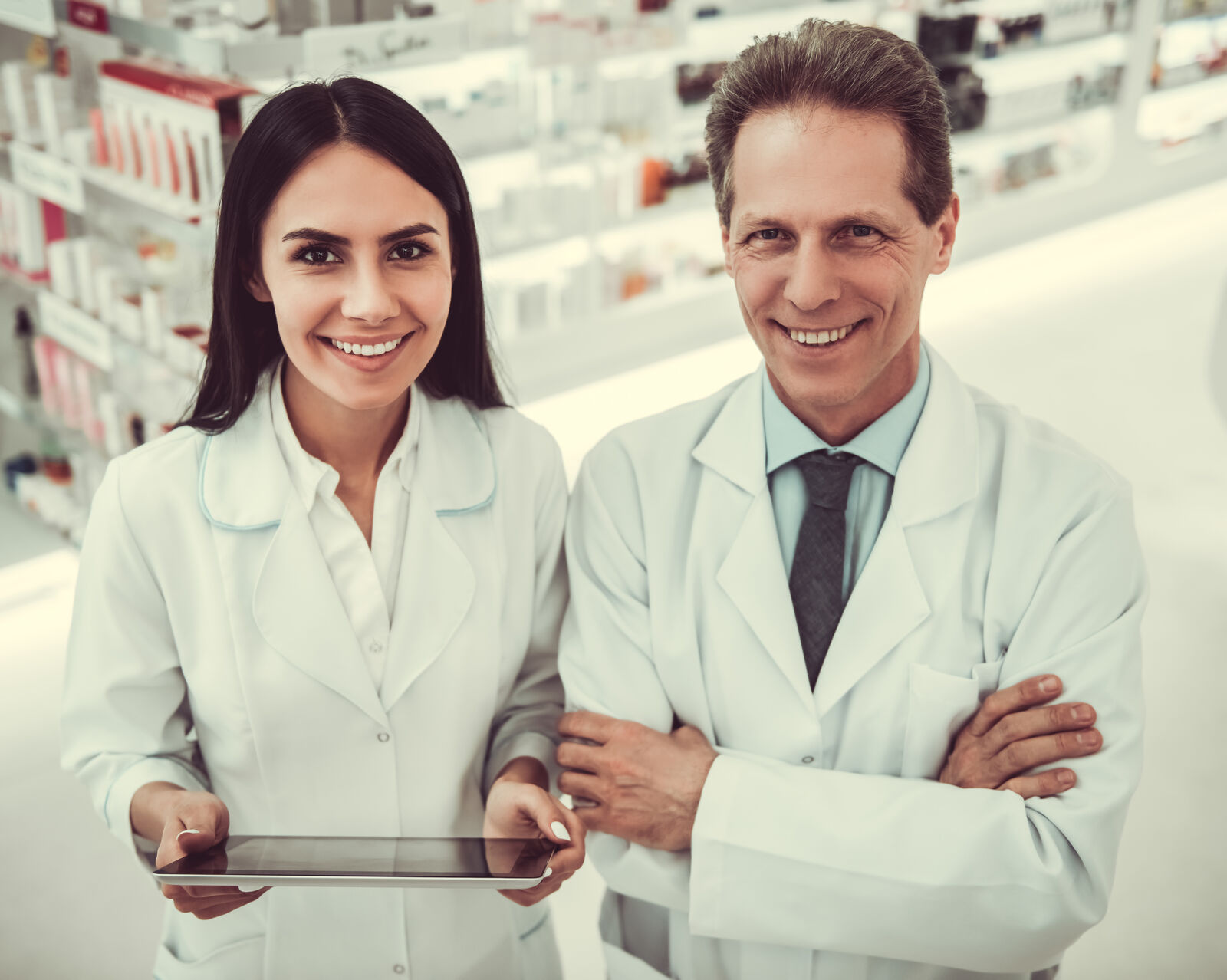 Nasmejani farmaceuti u belim mantilima stoje u apoteci, žena drži tablet, a muškarac prekrsti ruke.